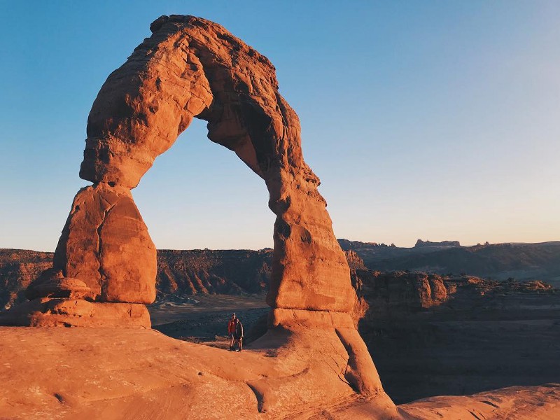 two people standing below a giant, orange, natural, stone arch