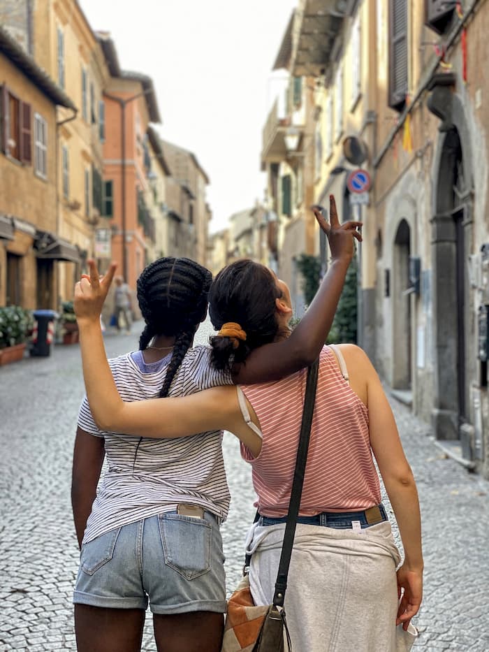 two teenage girls, black and white, with arms around each other's shoulders looking at buildings along a cobblestone street