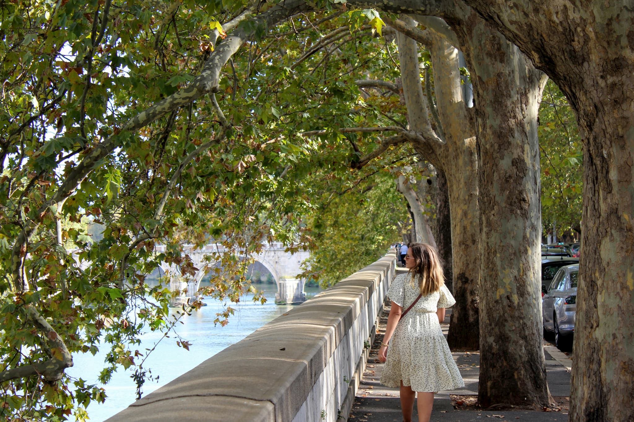 Woman walking down a sidewalk with tree branches stretching overhead towards the river down below