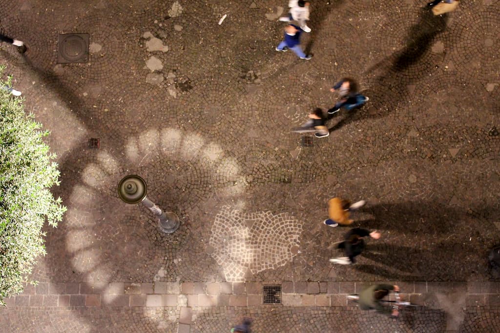 View of street from above with circular brick patterns, the light of a street lamp, and people blurred as they walk by.
