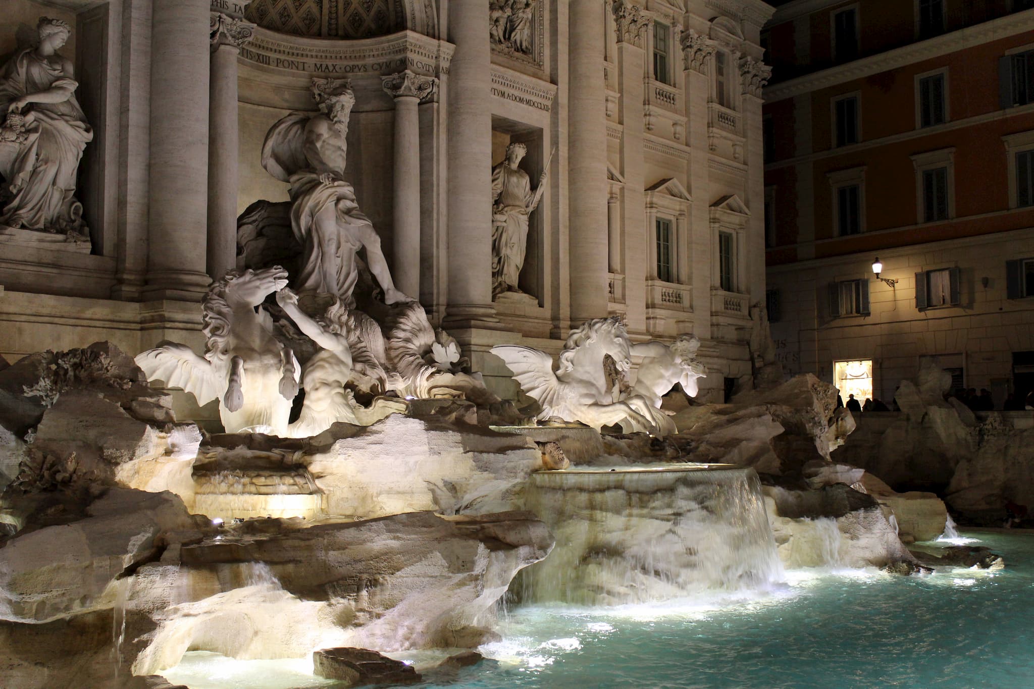 Marble carved statues forming a fountain behind a pool of water lit up in the nighttime