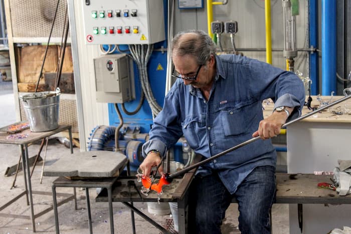 Man sitting, working a piece of hot glass with metal tools.