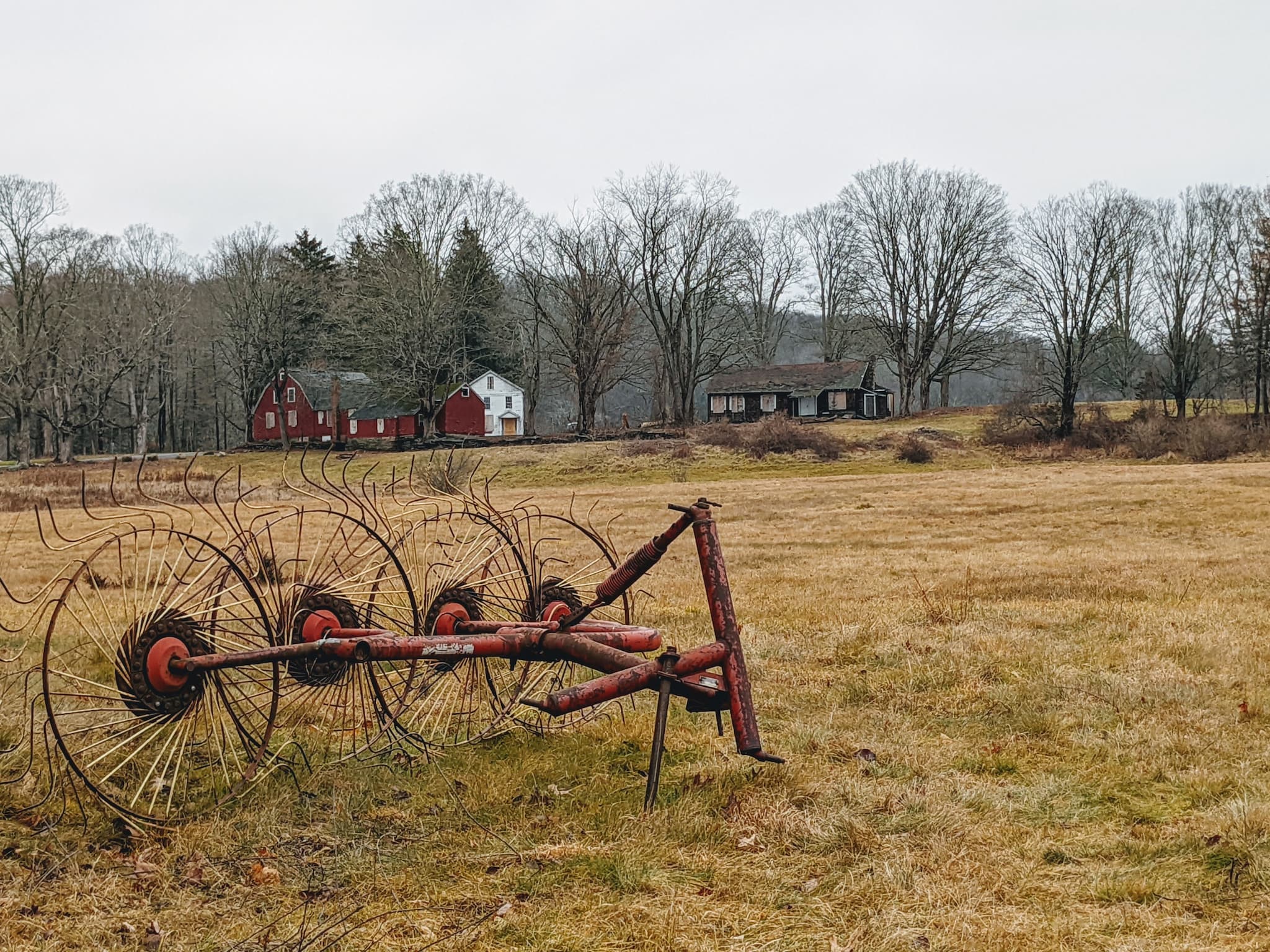 brown grass field with farm machinery in the foreground and buildings and bare trees in the background.