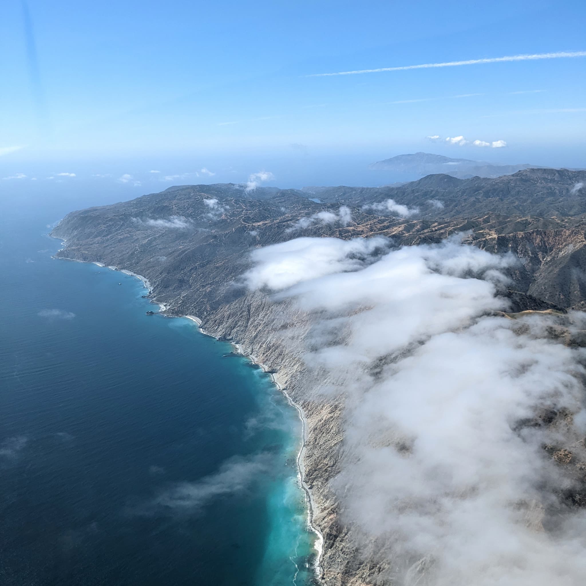 Looking down on deep blue waters along cliffs of a coastline with clouds below.