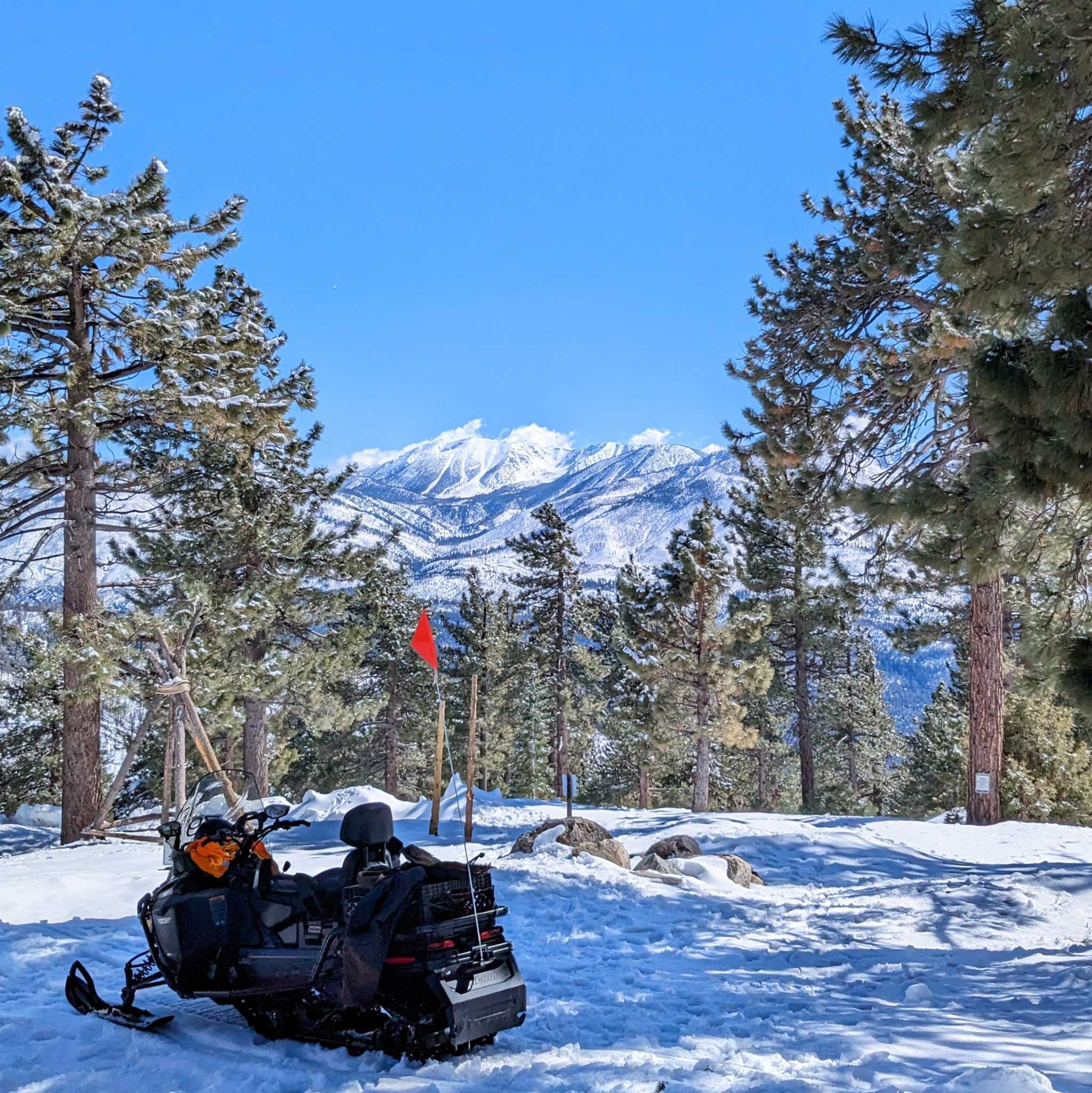 View of a snow covered mountain from between evergreen trees on an also snowy mountain top with a snowmobile.