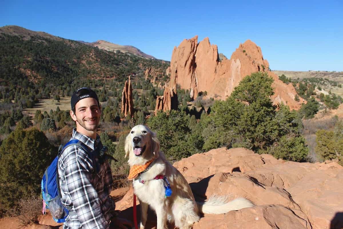 A man with a dog on top of a red, rocky bluff with hills and more red-rock spires behind.
