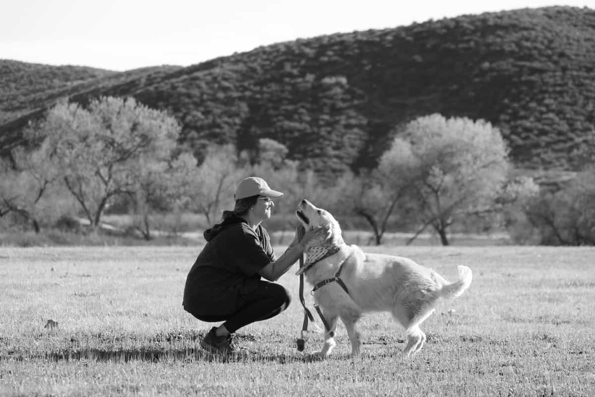 Black and white photo of a woman kneeling to pet a happy dog in a field with trees and hills behind.