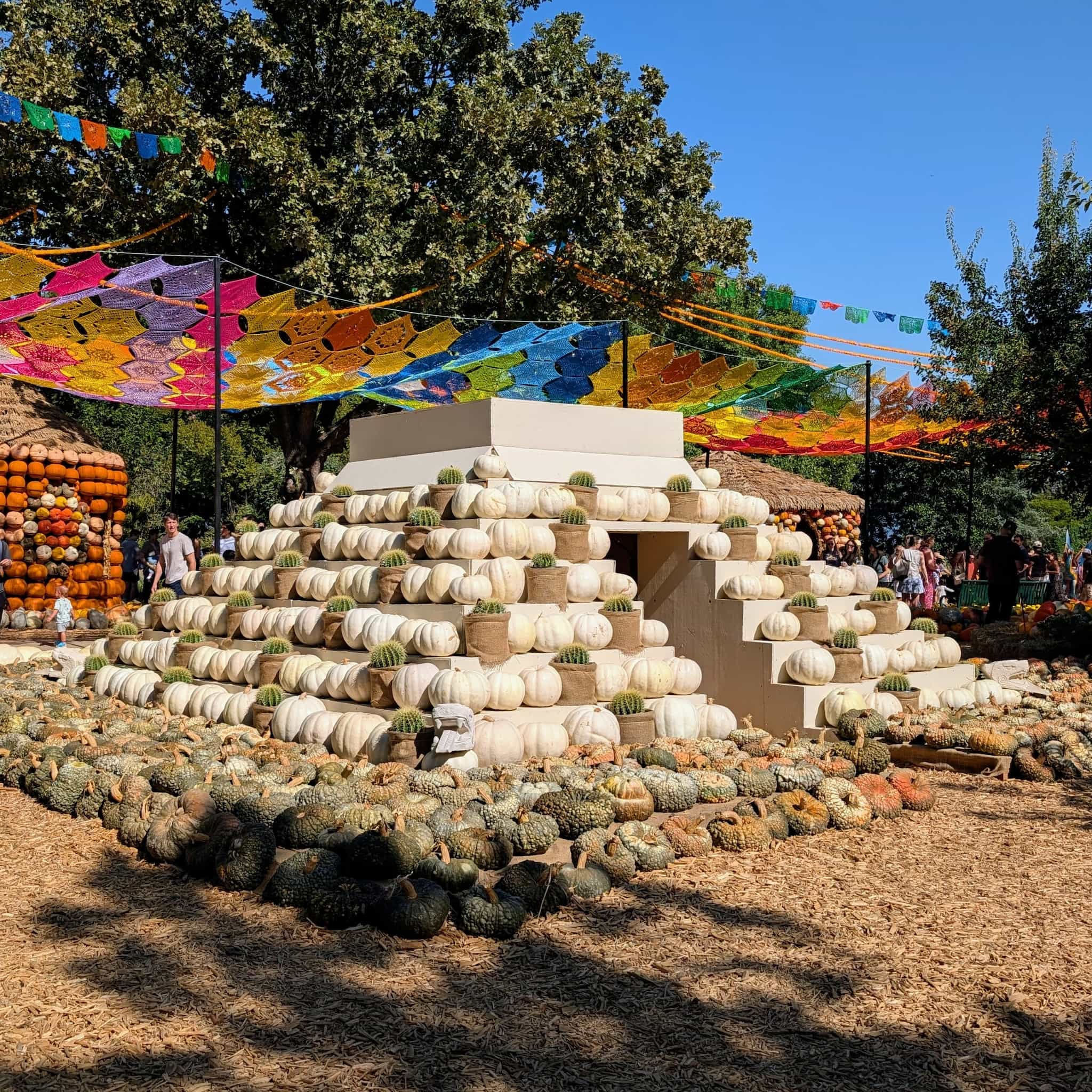 A white, stepped pyramid structure topped with white pumpkins and barrel cacti and a colorful Mexican cut-paper decoration behind.