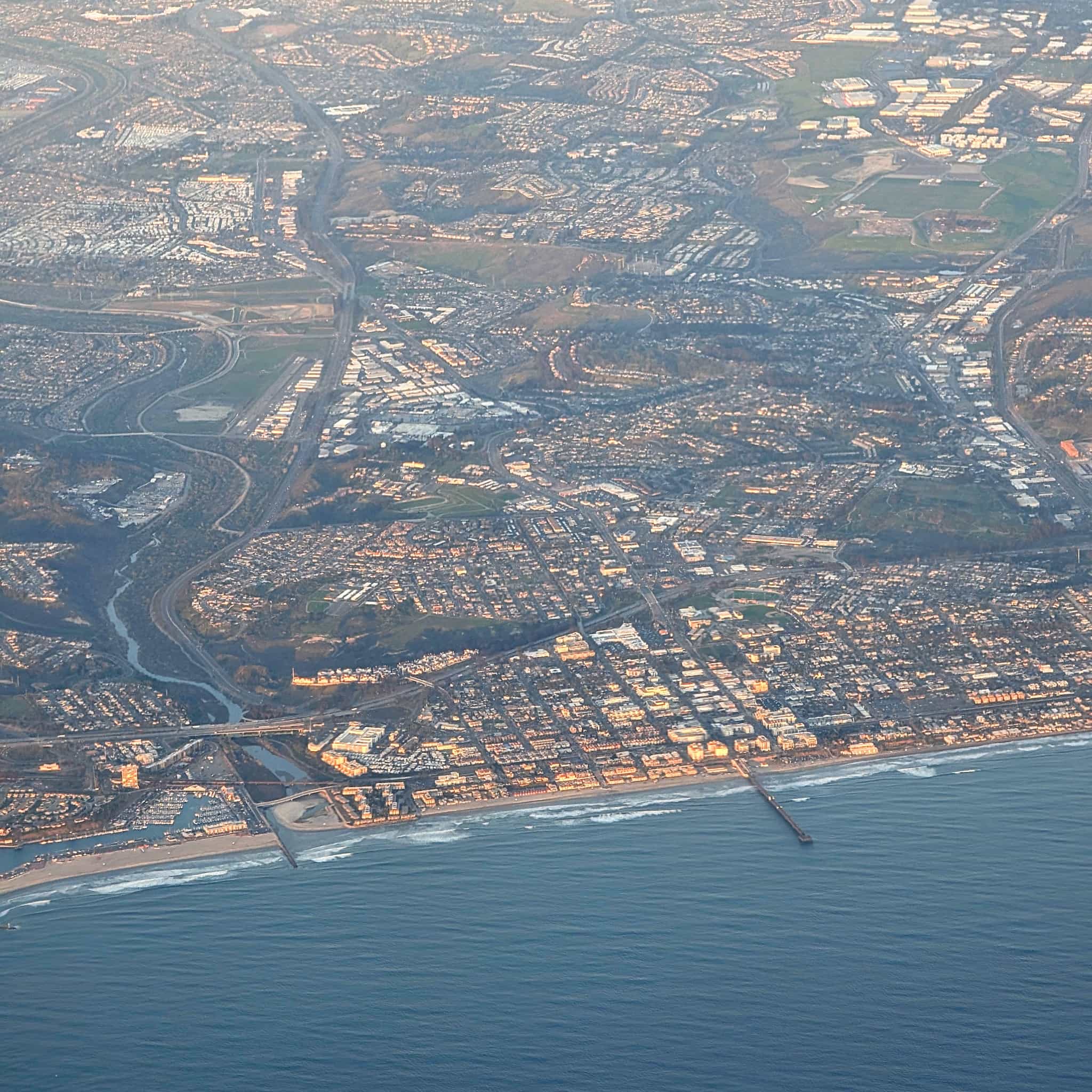 Areal photo of a straight coastline with buildings glowing orange with a straight pier sticking out into the ocean and a river flowing from deeper inland.