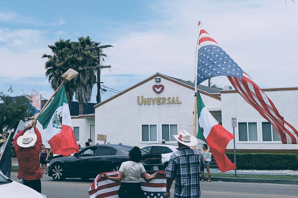 Protesters holding upside down American flags and waving a Mexican flag tied to a shovel in front of a church building with a sign reading 'Universal Love'
