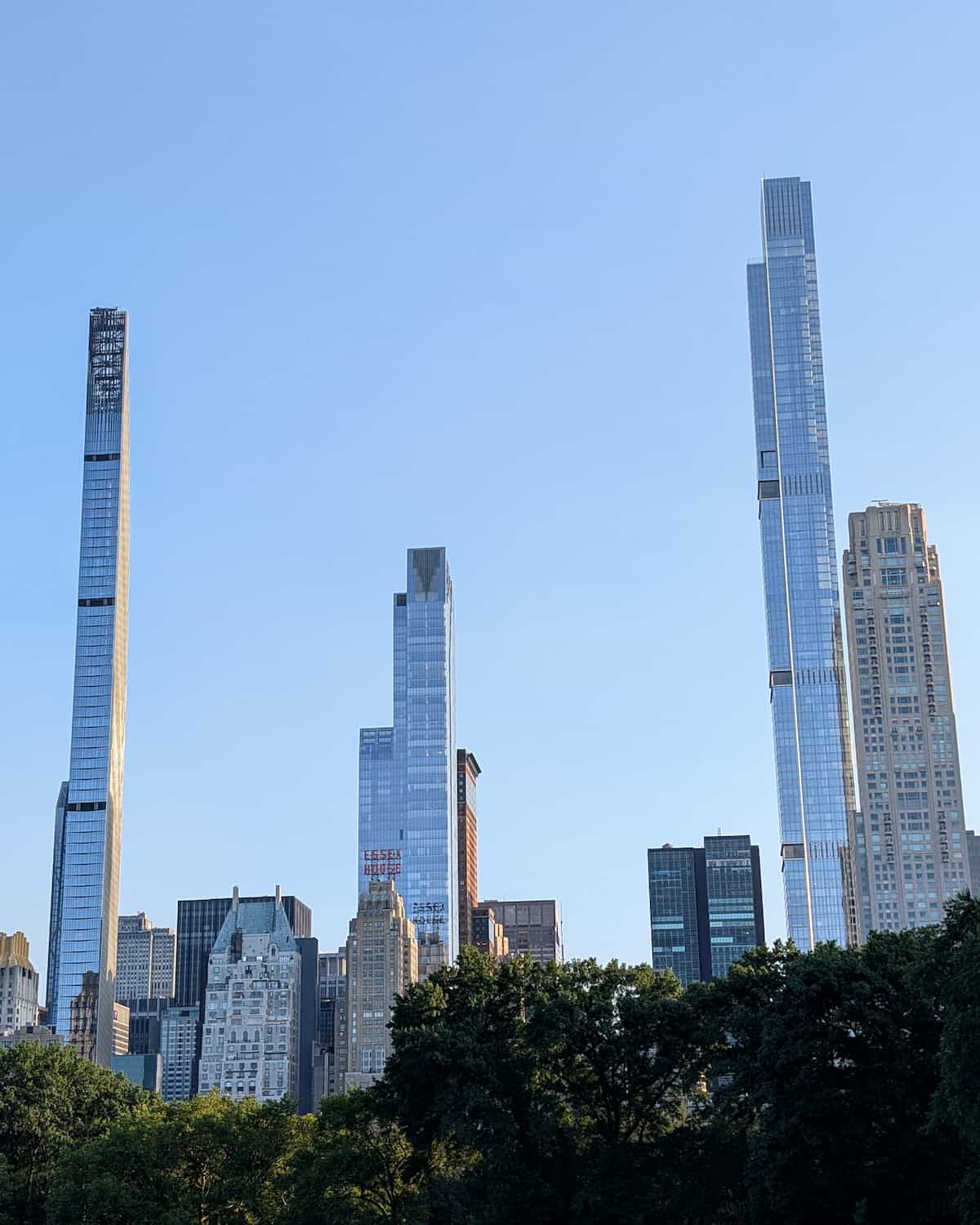 Four sky scrapers reaching into a blue sky with shorter buildings and treetops below.