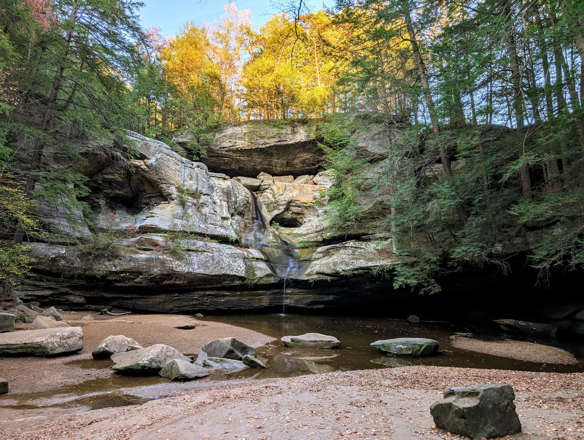 A large cliff face with a small amount of water dribbling down framed by trees with golden leaves on top, green trees on the side, and a pool of water below.