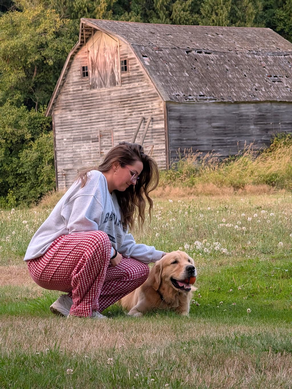 A woman and golden retriever sitting in a field in front of a barn.
