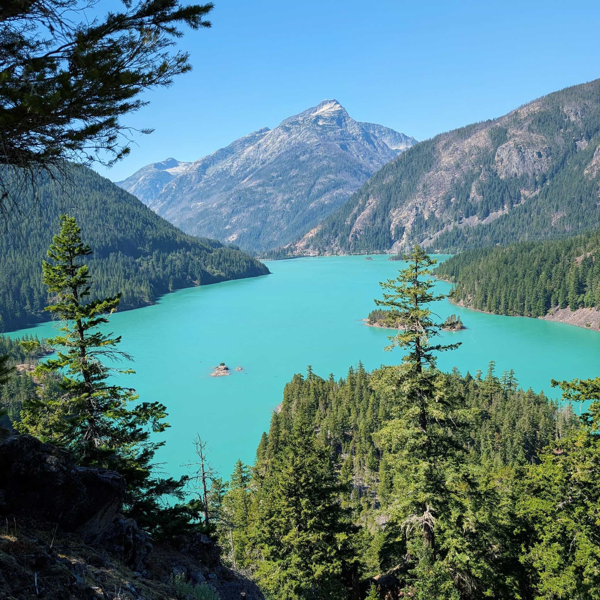 Looking down on a light blue lake surrounded on all sides by evergreen-covered slopes with a mountain peak visible in the distance.