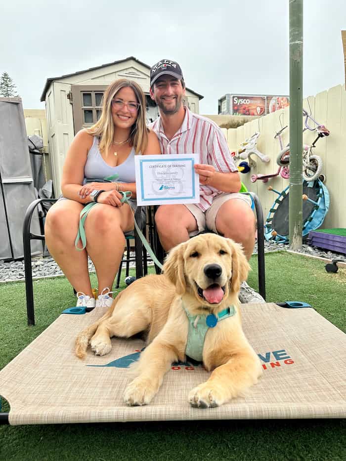 Two people sit behind a dog facing the camera and holding a certificate