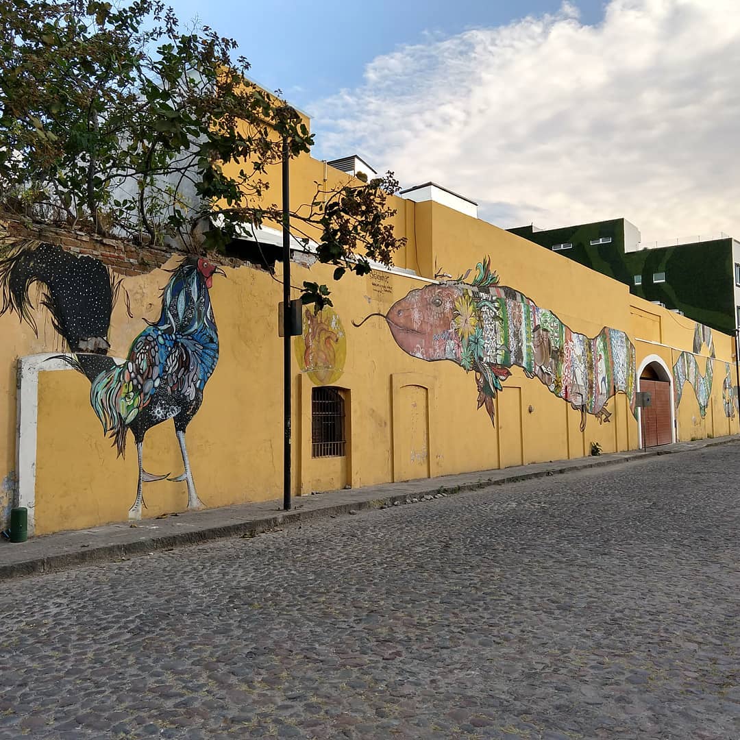 Long yellow building on an empty cobblestone street. The building has murals of a chicken and a long dragon-like creature.