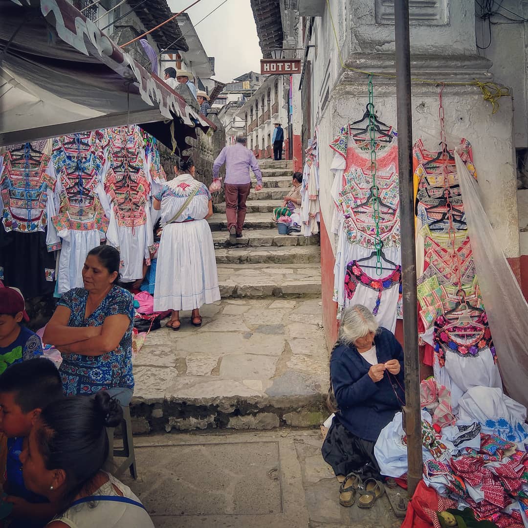 Stone steps in a mountain village path. One building has a sign that reads 'hotel'. Women stand in front of displays of white embroidered shirts.