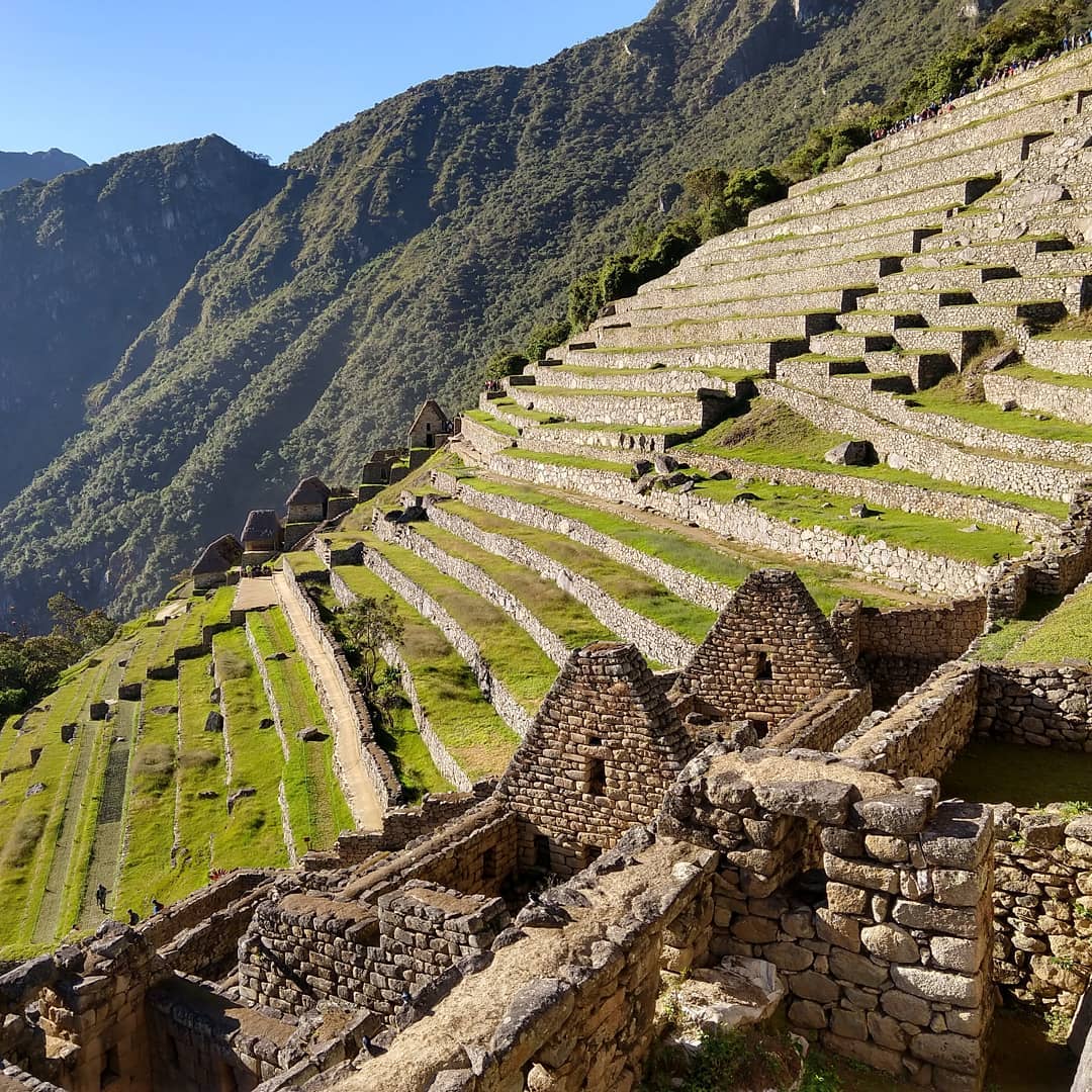 Steep mountainside terraced into flat grassy areas using stone walls. The foundation of stone buildings is visible in the foreground.