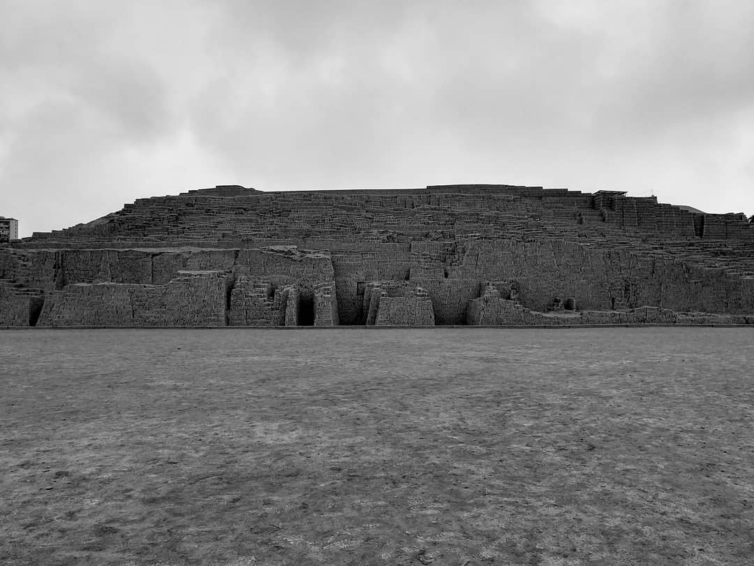 Grayscale image of an uneven brick mound with a large sandy area in front and a cloudy sky above.