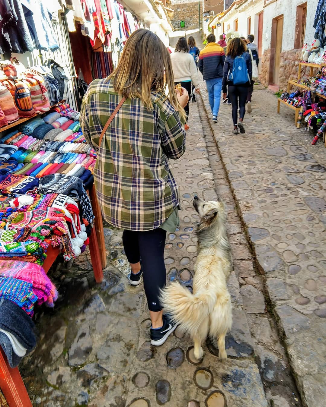 Woman looking down at a dog that is looking up at her, walking down a narrow cobblestone street where colorful clothes are for sale.