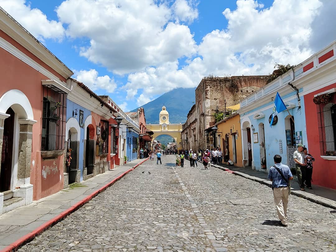 Cobblestone street on a sunny day with colorful buildings and a yellow arch at the end with mountains in the distance.