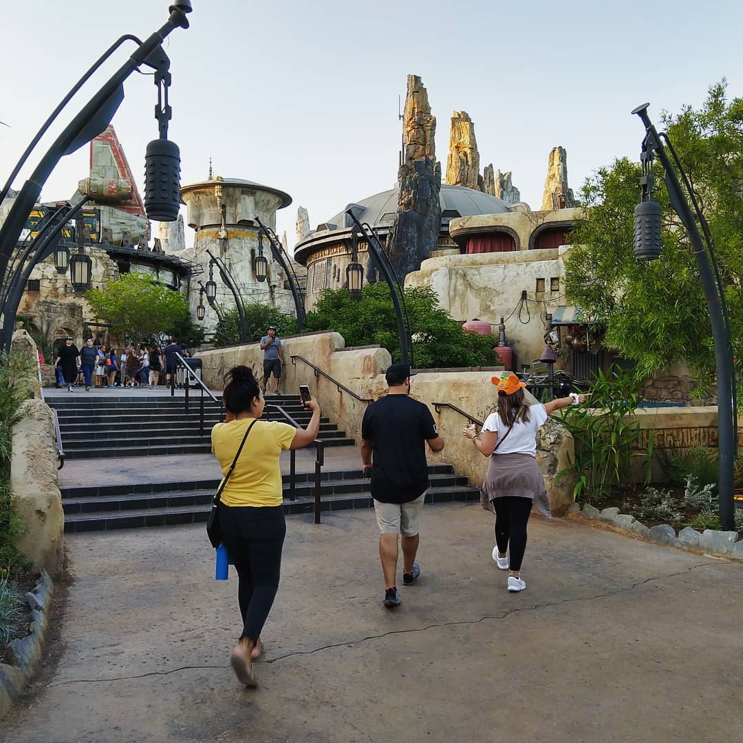 Three adults walking in a Star Wars themed adventure land. One is pointing to the side. Another with walking around recording with a cell phone.