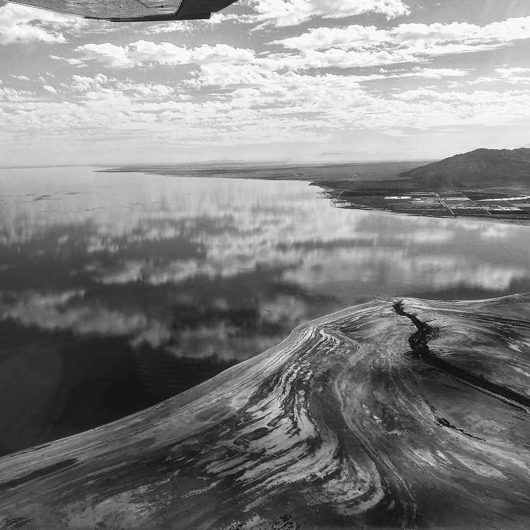 Black and white photo looking down on a large body of water with clouds in the reflection. A stream flows through a bare and muddy shoreline.