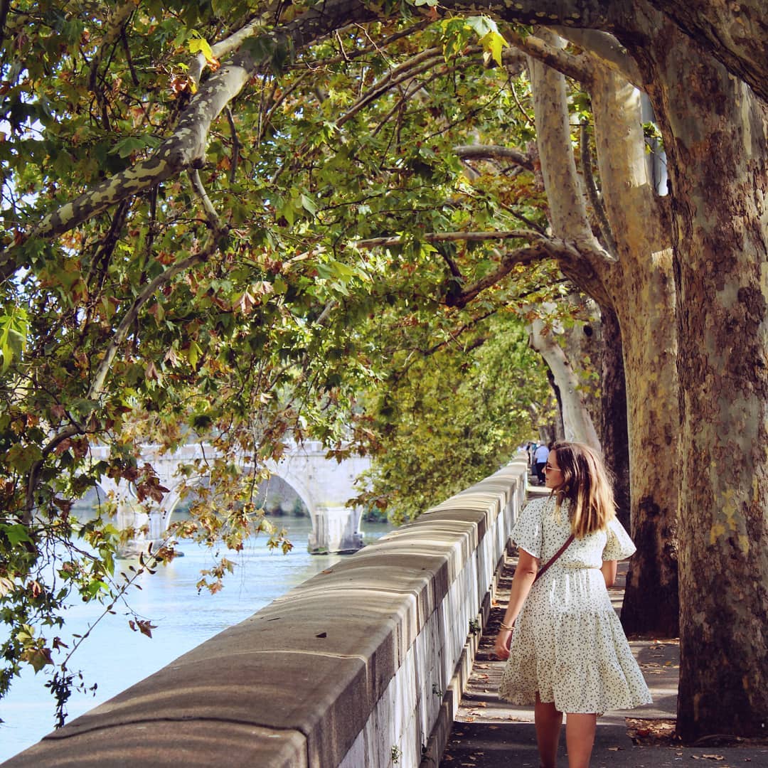 A woman walking down a tree-linded sidewalk overlooking a river.