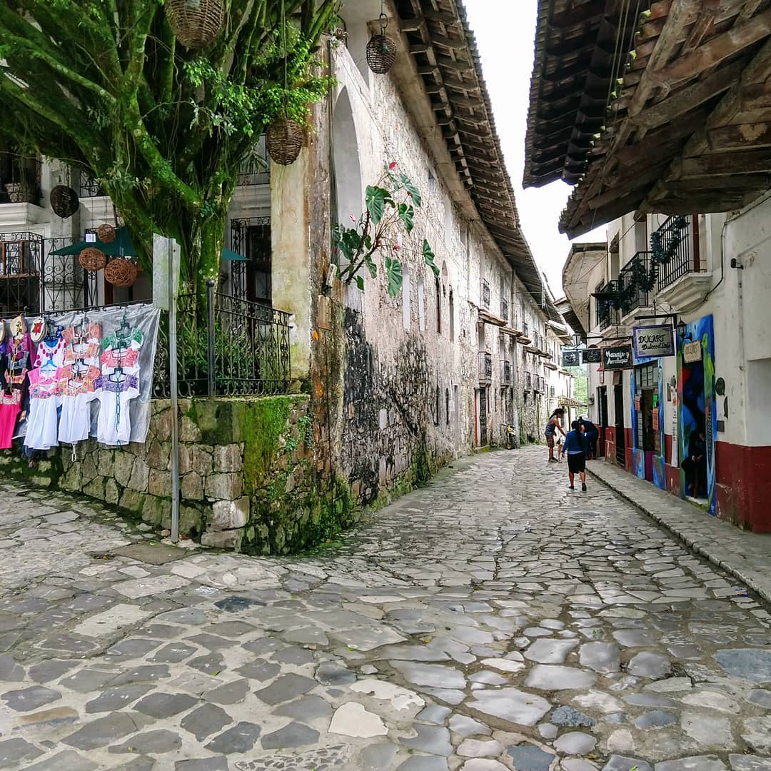 Rustic cobblestone street with green plants, colorful shirts, and signs in shop windows.