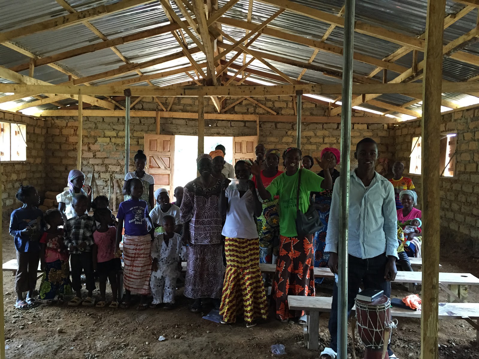 inside a mud brick building with about 15 adults in colorful dress and 10 kids standing next to wooden benches