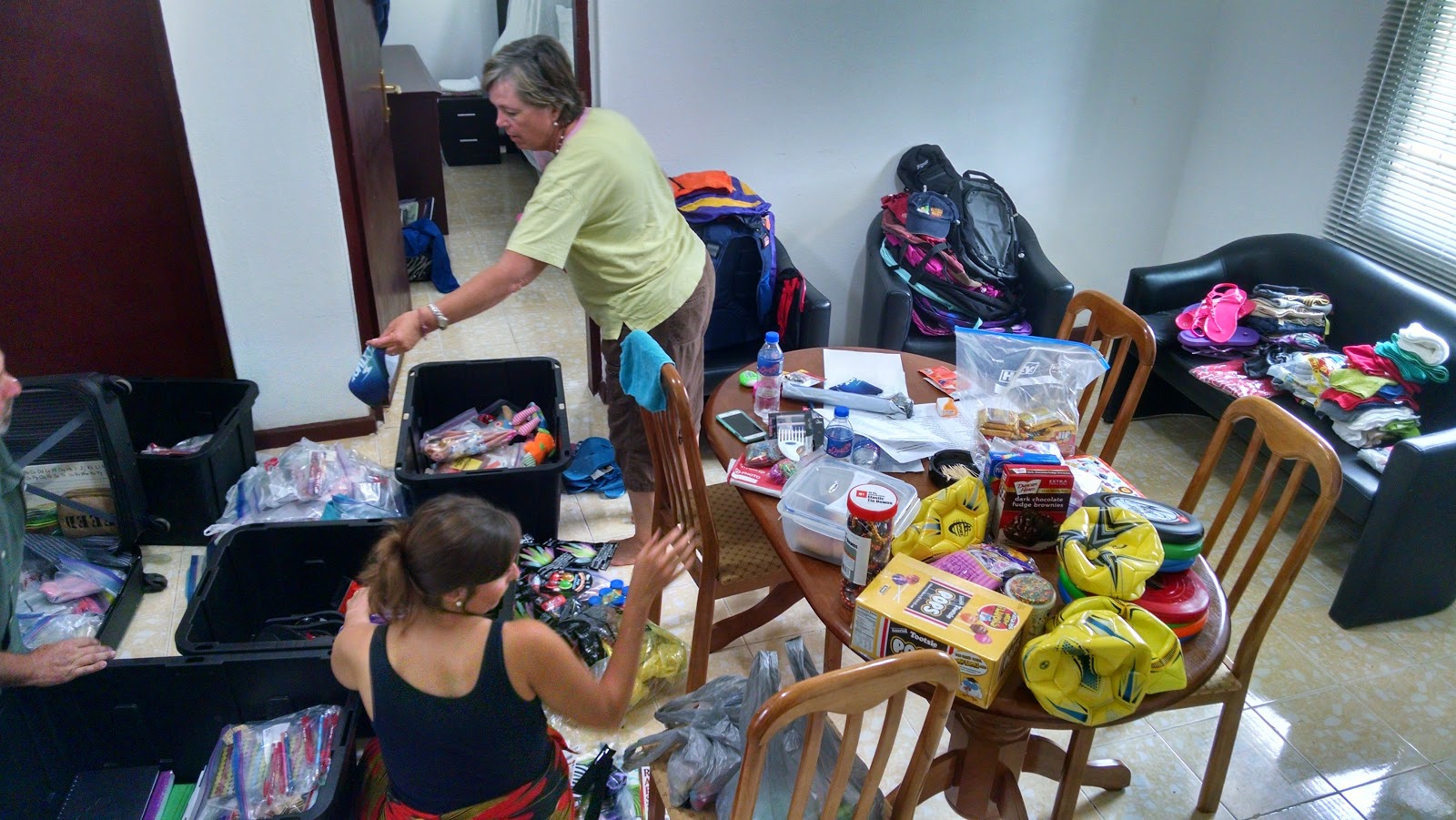 room with toys and other donations laid across tables and couches with open suitcases on the floor and two people organizing the items