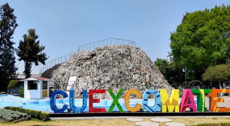 Colorful letters mounted to the ground spelling Cuexcomate with a rocky mound about 30 feet tall in the background with stairs to the top