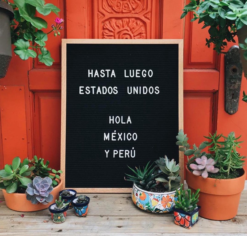 black letter board with white letters reading Hasta luego estados unidos, hola mexico y peru with plants around it