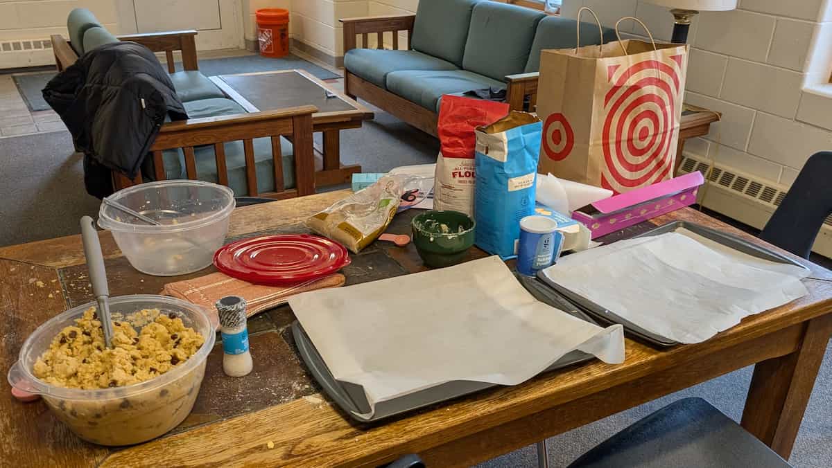 Messy table with baking sheets, tupperware containers, baking ingredients, and a Target bag in a sloppy dorm common room.