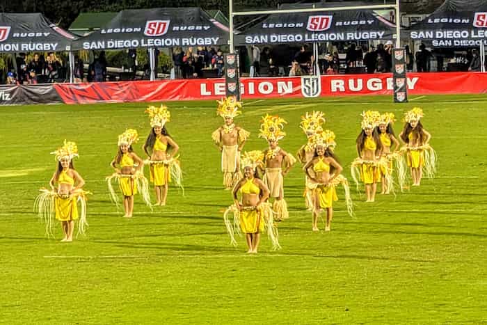 Men and women in traditional Polynesian dress in organized lines on the rugby pitch for a dance routine.