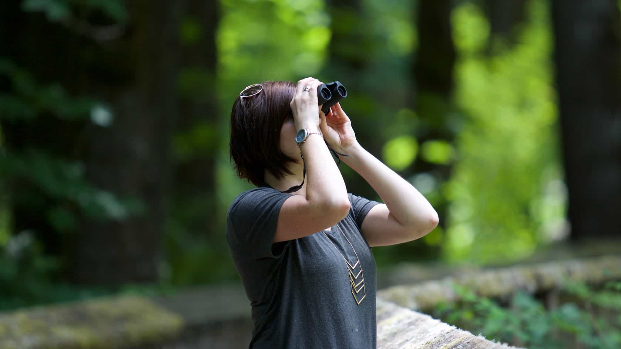Woman looking up at trees through binoculars.