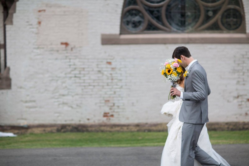 Couple on wedding day walking outside with flowers, faces obscured, wall of church in background